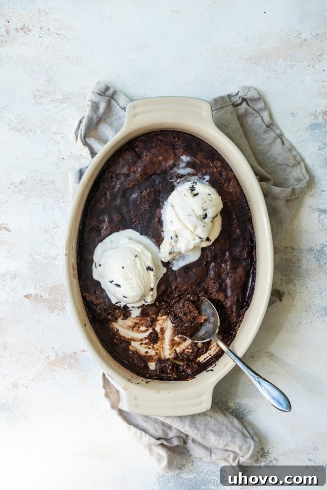 An aerial view of the freshly baked Brownie Chocolate Pudding Cake, still in its elegant oval baking dish, generously adorned with scoops of melting vanilla ice cream, creating a mouth-watering scene.