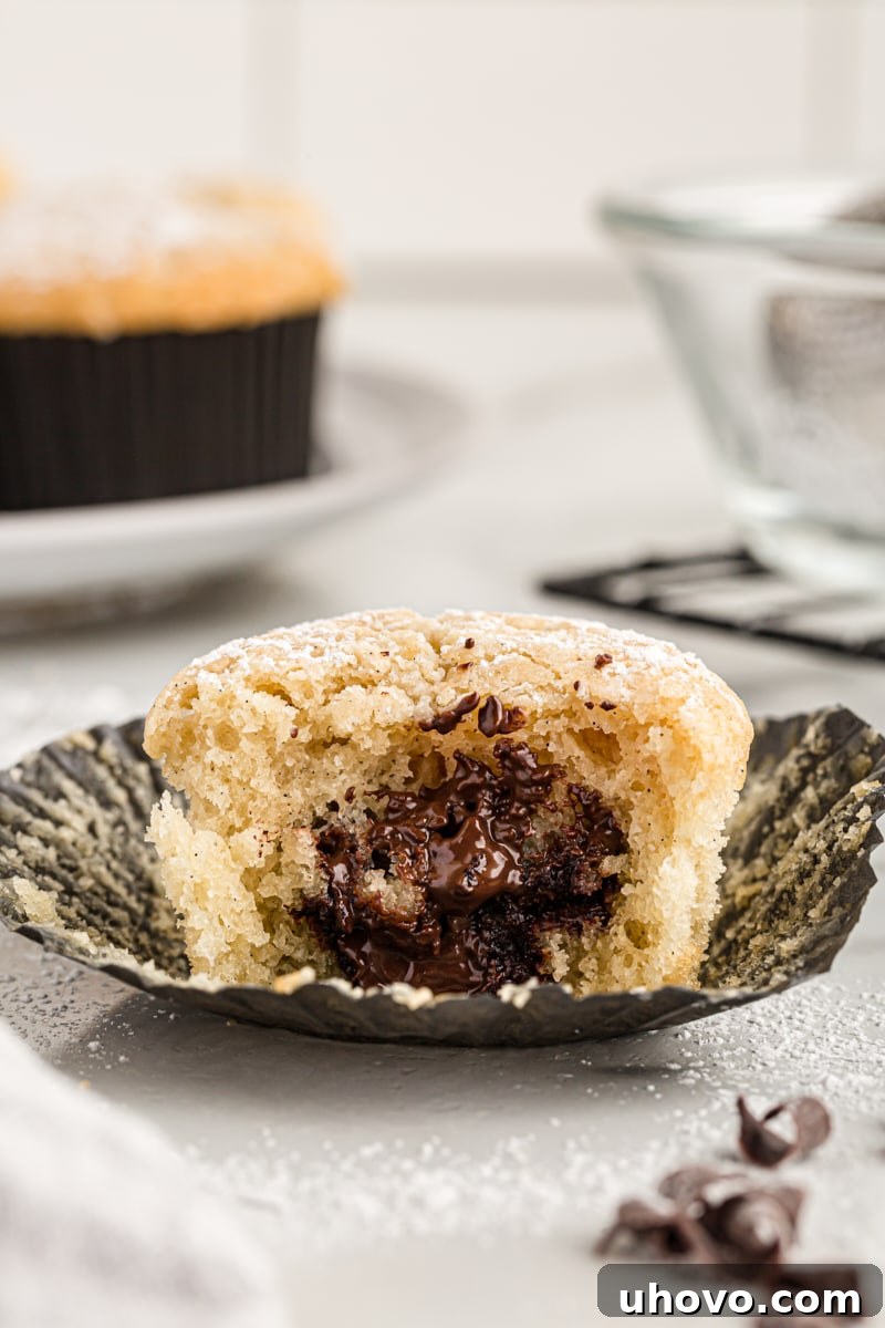 A chocolate-filled vanilla cupcake with a bite taken out, revealing the melted dark chocolate core, dusted with powdered sugar.
