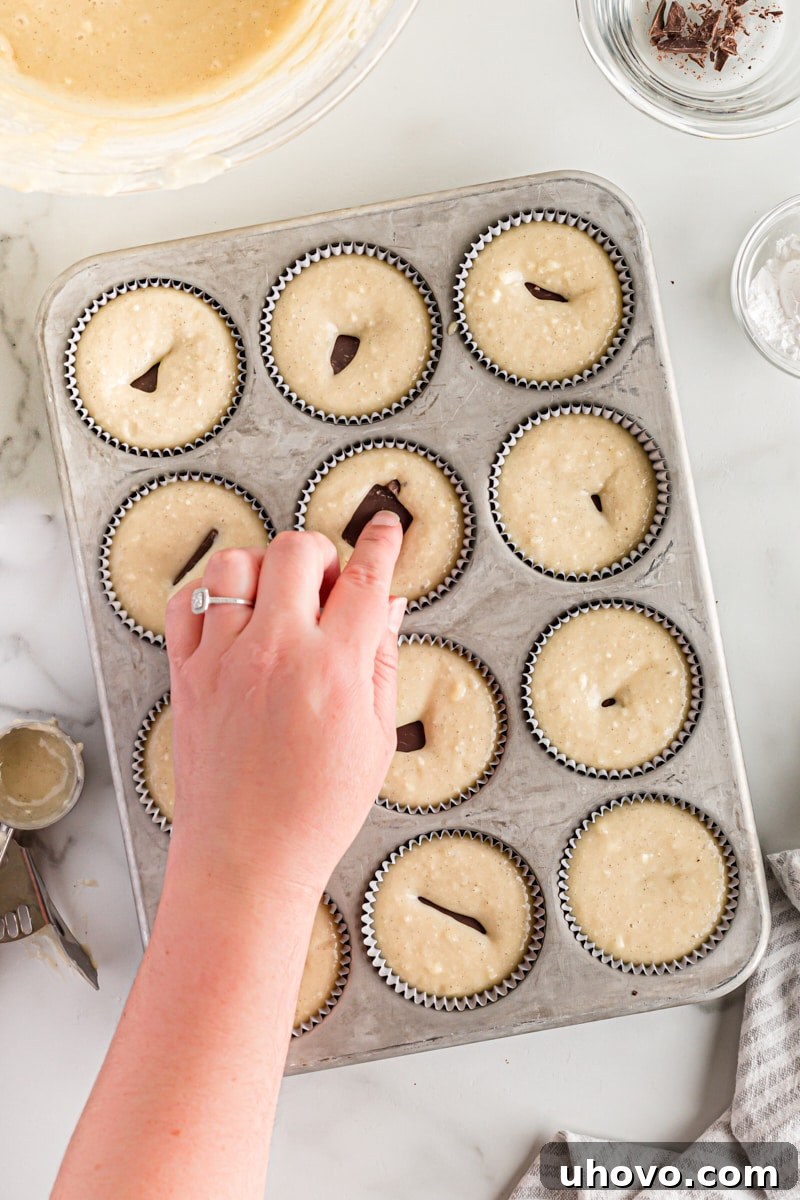 A hand gently pressing a piece of bittersweet chocolate into the center of cupcake batter in a muffin tin.