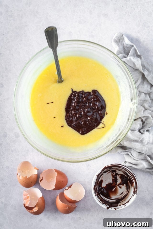 overhead view of melted chocolate being gently added to a creamy mixture of butter, sugar, and eggs in a glass mixing bowl
