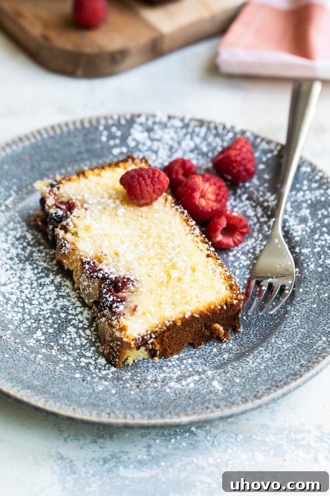 Slice of almond loaf dusted with powdered sugar on a plate with fresh raspberries and a fork, ready to be enjoyed.