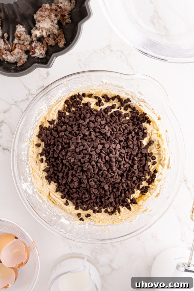 An overhead view of mini chocolate chips being added to the smooth chocolate chip Bundt cake batter in a glass mixing bowl.