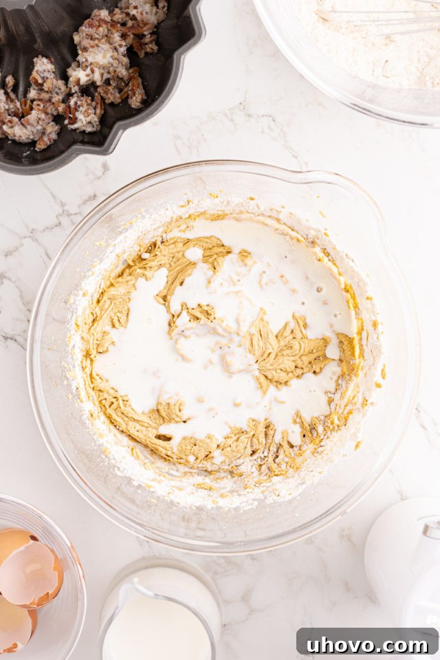 Buttermilk being gently poured into the chocolate chip Bundt cake batter in a glass mixing bowl, continuing the alternation of wet and dry ingredients.