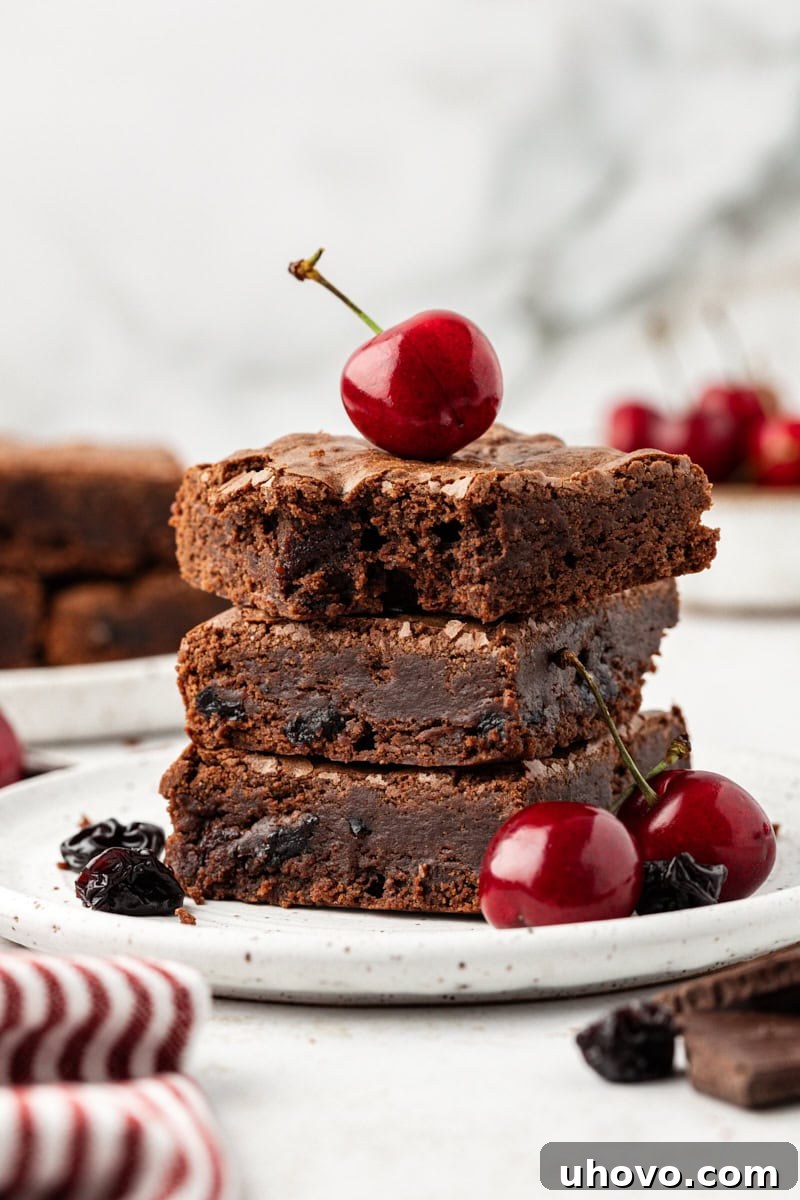 Three dark chocolate sour cherry brownies stacked on plate, showcasing their rich, fudgy texture and bits of cherry.