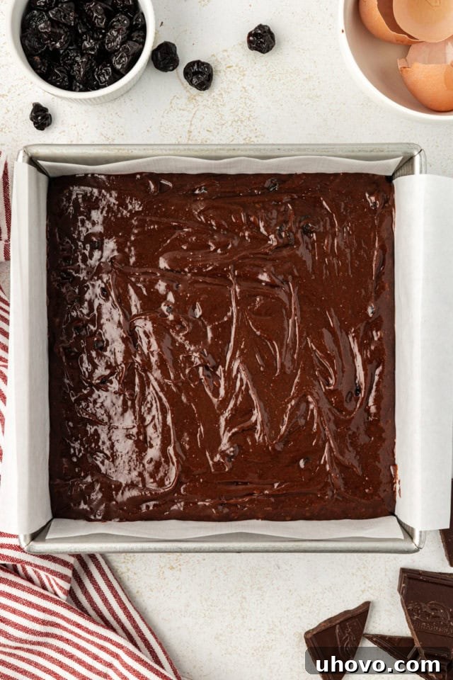 Dark chocolate sour cherry brownie batter spread evenly in a parchment-lined 8-inch square baking pan, ready for the oven.