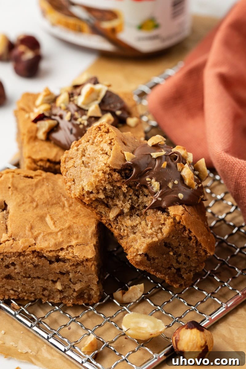 A hazelnut blondie with a bite taken out, leaning against two more perfectly baked blondies on a wire cooling rack, inviting a taste.