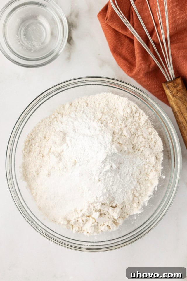 Overhead view of all-purpose flour, baking powder, and salt meticulously measured and combined in a clear glass mixing bowl, ready for the next step in making hazelnut blondies.