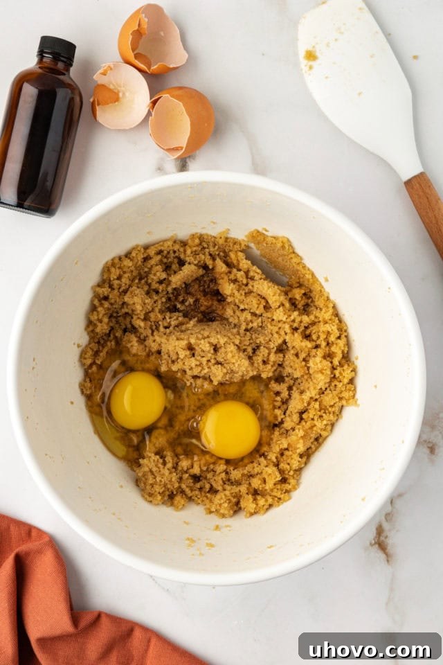 A close-up, overhead view showing large eggs and a splash of vanilla extract being carefully added to a bowl containing the perfectly combined mixture of melted butter and light brown sugar.