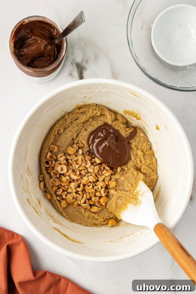 An overhead view of chopped toasted hazelnuts and a dollop of chocolate-hazelnut spread being carefully folded into the rich blondie batter in a mixing bowl.