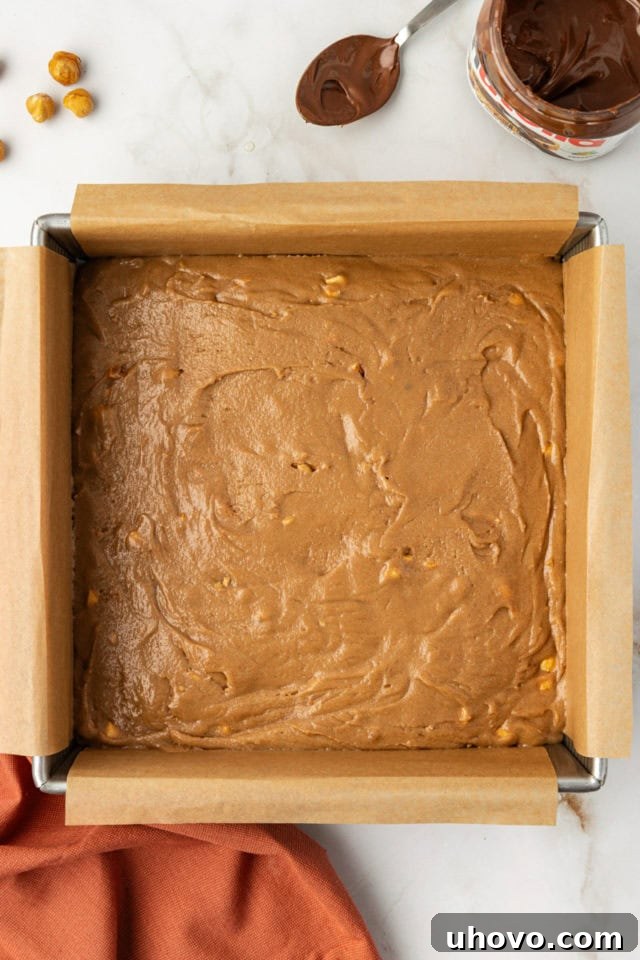 A smooth, golden hazelnut blondie batter evenly spread across the bottom of a square baking pan, ready for the oven.