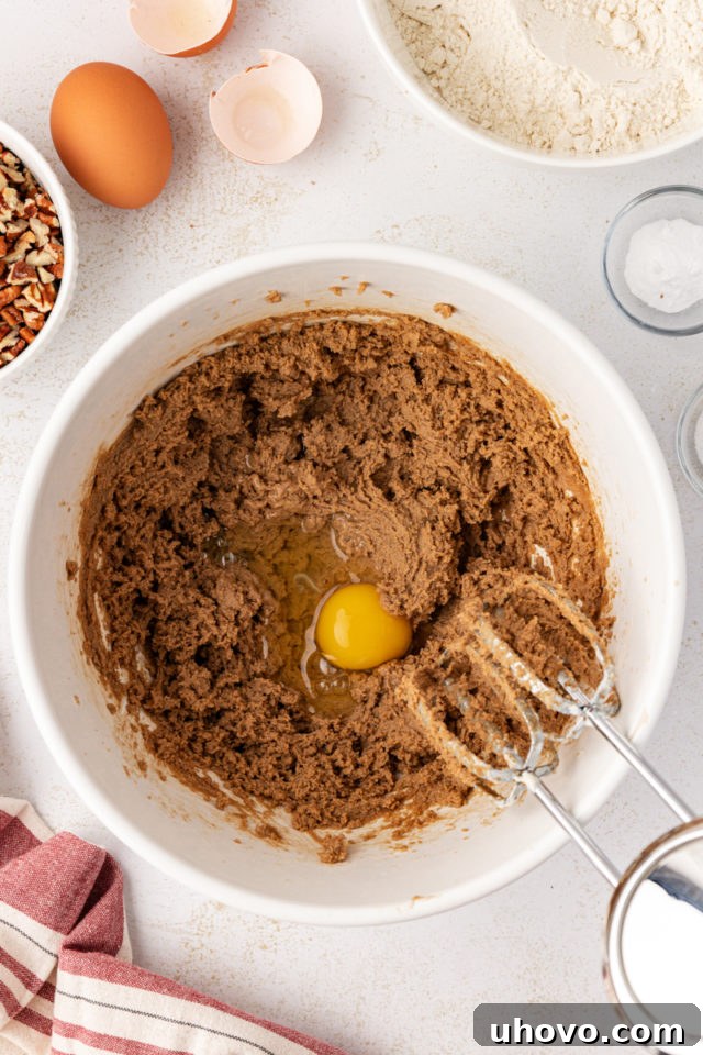 Egg being added to a creamy mixture of brown sugar, butter, and vanilla in a mixing bowl.