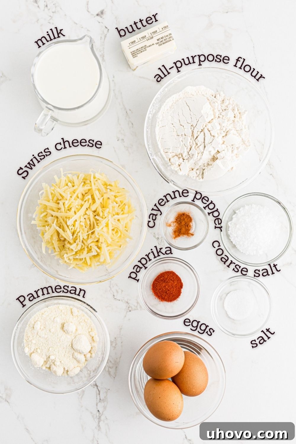 Overhead view of all the ingredients laid out on a table for making gougères, including milk, butter, flour, eggs, and various cheeses.