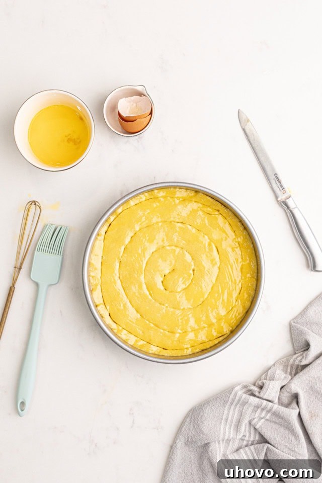 A baker's hand gently scoring a traditional crisscross pattern onto the egg-washed surface of the almond gâteau Breton batter in a cake pan, just before baking.
