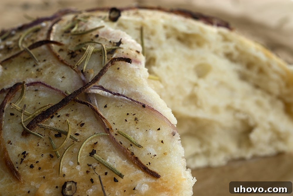 Golden focaccia bread, topped with thinly sliced onions and fresh rosemary, rests on a wooden cutting board, ready to be served. Its crust is perfectly browned, hinting at a soft, aromatic interior.