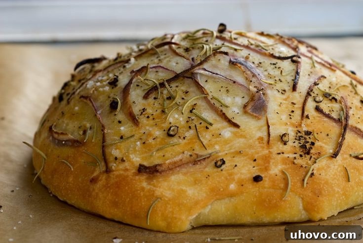 Golden focaccia bread, topped with thinly sliced onions and fresh rosemary, rests on a wooden cutting board, ready to be served. Its crust is perfectly browned, hinting at a soft, aromatic interior.