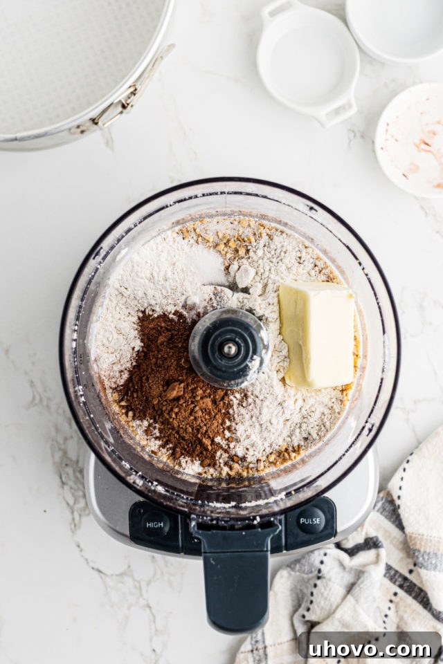 Overhead view of all remaining crust ingredients – flour, butter, cocoa powder, and salt – added to the food processor with the chopped hazelnuts.