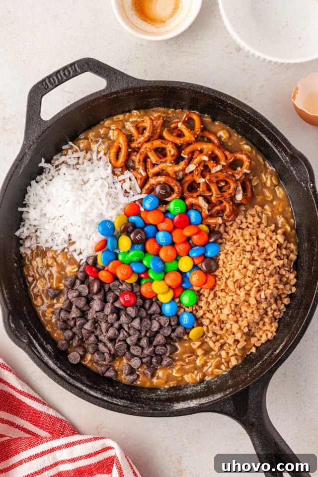 overhead view of chocolate chips, M&Ms, pretzels, and more add-ins added to cookie dough in a cast iron skillet