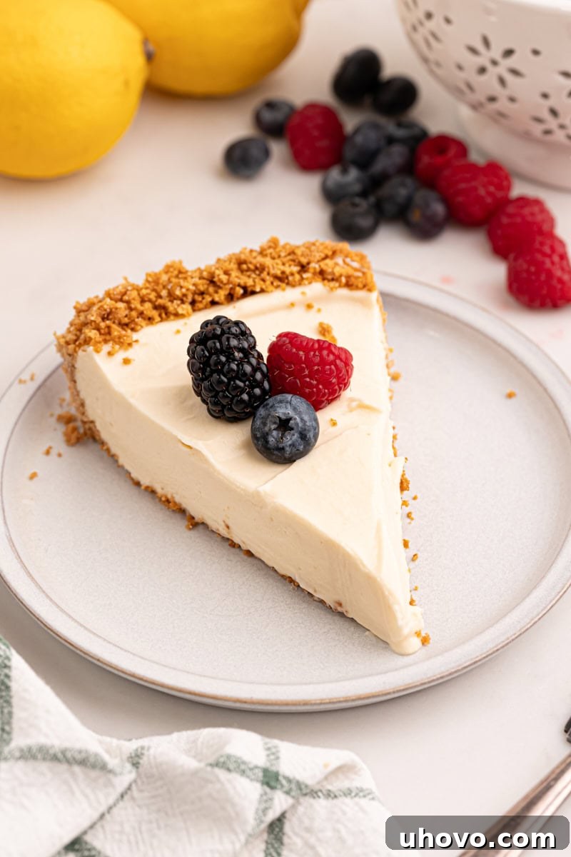 A beautifully presented slice of lemon cream pie, garnished with an array of fresh, vibrant berries and a delicate sprig of mint. In the softly blurred background, an entire lemon cream pie sits alongside additional fresh berries and two bright yellow lemons, highlighting the dessert's natural, zesty ingredients and refreshing appeal.