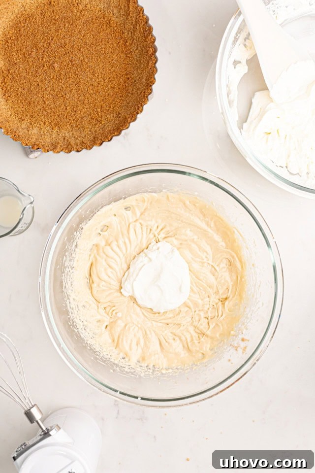 An overhead view of a clear glass mixing bowl where a portion of whipped cream has just been added to the lemon cream cheese mixture. The process of gently folding them together is visible, aiming for a light and airy consistency.