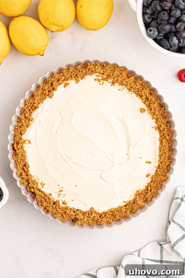 An overhead view of a finished lemon cream pie, perfectly chilled in its pan, surrounded by whole fresh lemons and a small bowl of mixed berries. The pie's smooth, pale yellow surface is ready for garnishing and serving, exuding freshness and appeal.