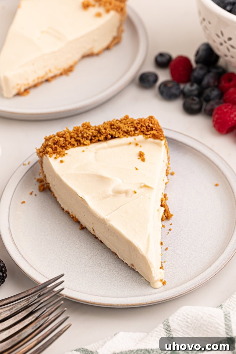 A close-up of a single slice of lemon cream pie elegantly placed on a white plate, topped with a vibrant medley of fresh berries. In the background, another slice of pie and a bowl brimming with more berries are softly blurred, creating a tempting and fresh dessert scene.