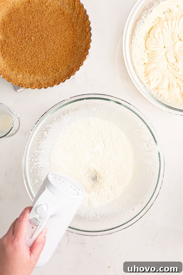 An electric mixer with a whisk attachment actively whipping cold heavy cream in a clear glass mixing bowl. The cream is starting to thicken and show visible peaks, indicating the progress of whipping.