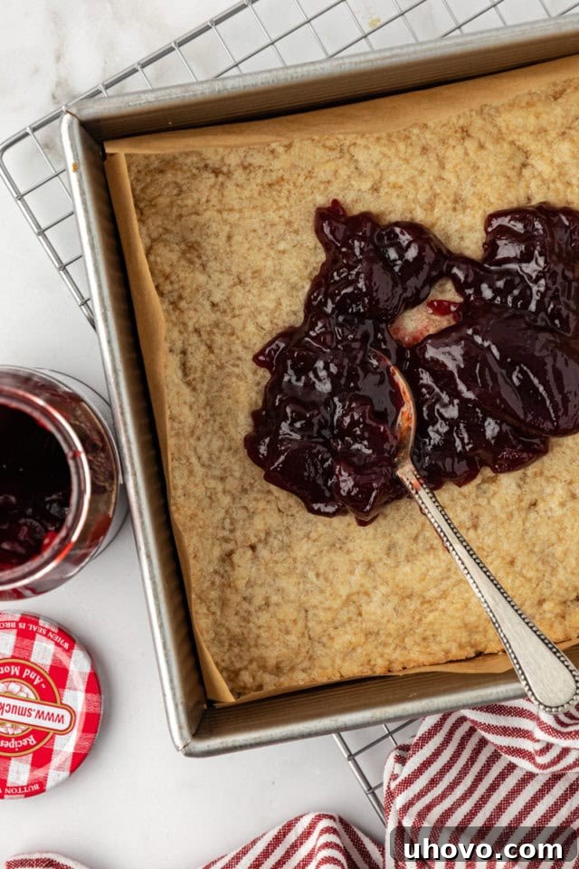 Overhead view of raspberry preserves being spread evenly onto the baked crust with a spoon