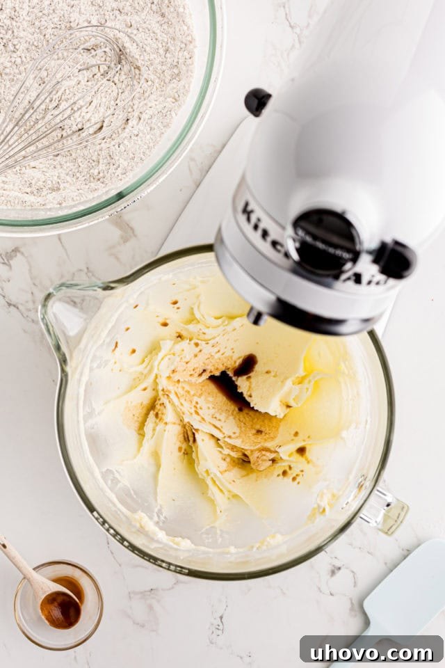 Vanilla extract being drizzled into a stand mixer bowl containing creamed butter, cream cheese, and sugar.