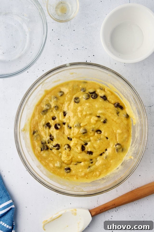 Close-up overhead view of the finished chocolate chip muffin batter in a glass mixing bowl, showing a slightly lumpy texture with visible streaks of flour, indicating perfect mixing.