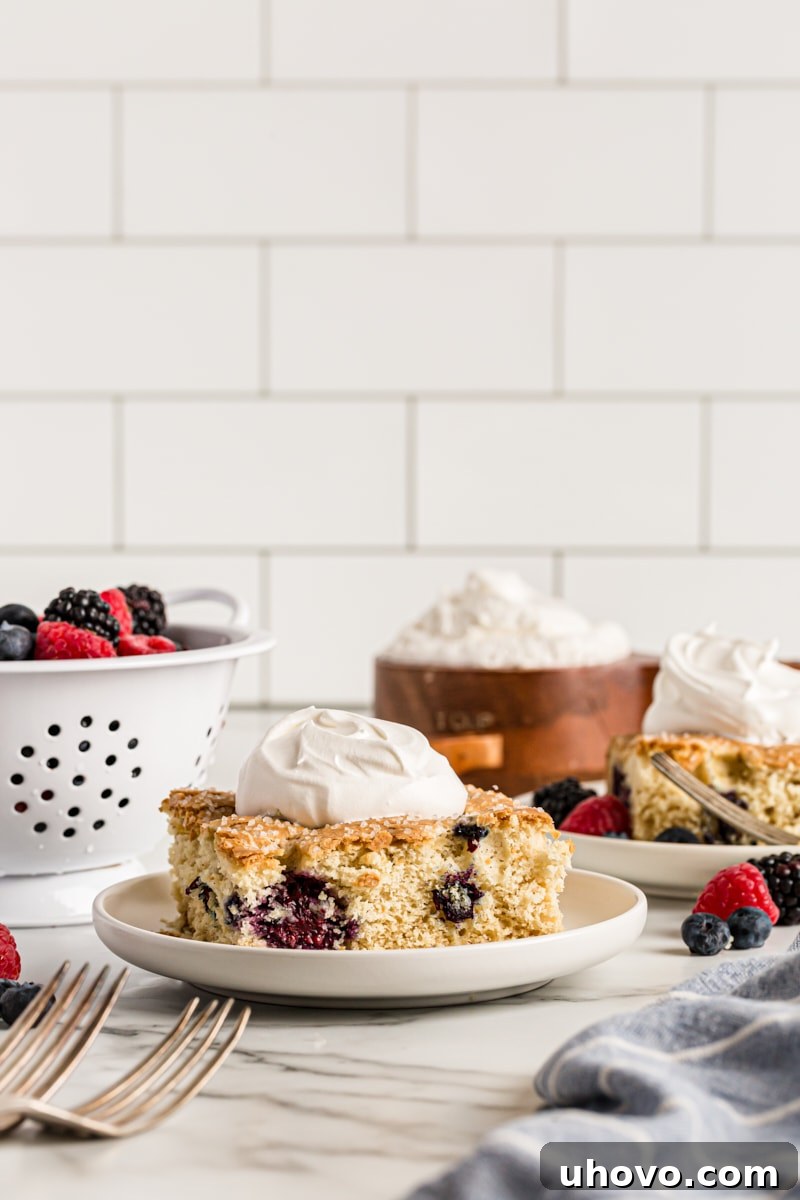 Square of berry vanilla cake on plate with dollop of whipped cream, berries, whipped cream, and another plate in background