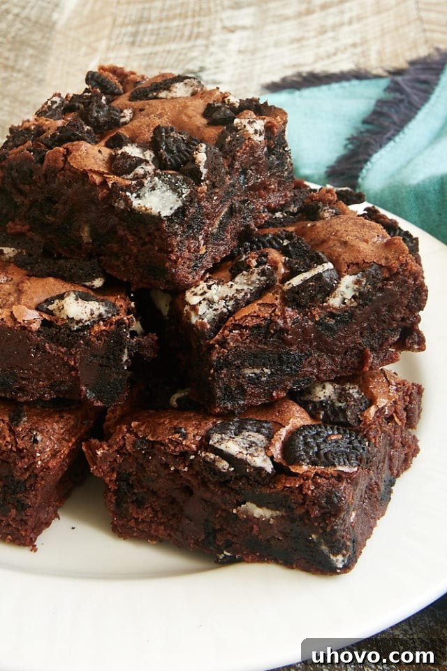 Stack of freshly baked Fudgy Cookies and Cream Brownies on a pristine white plate, showcasing their rich, dark chocolate interior and visible cookie pieces.