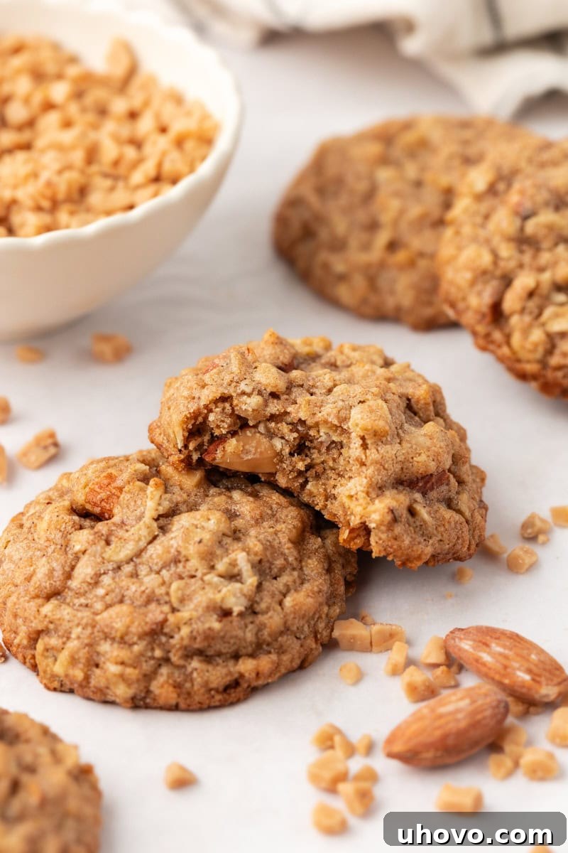 Two golden Toffee Almond Oatmeal Cookies, one with a bite taken out, showcasing the chewy texture and visible toffee and almond pieces.