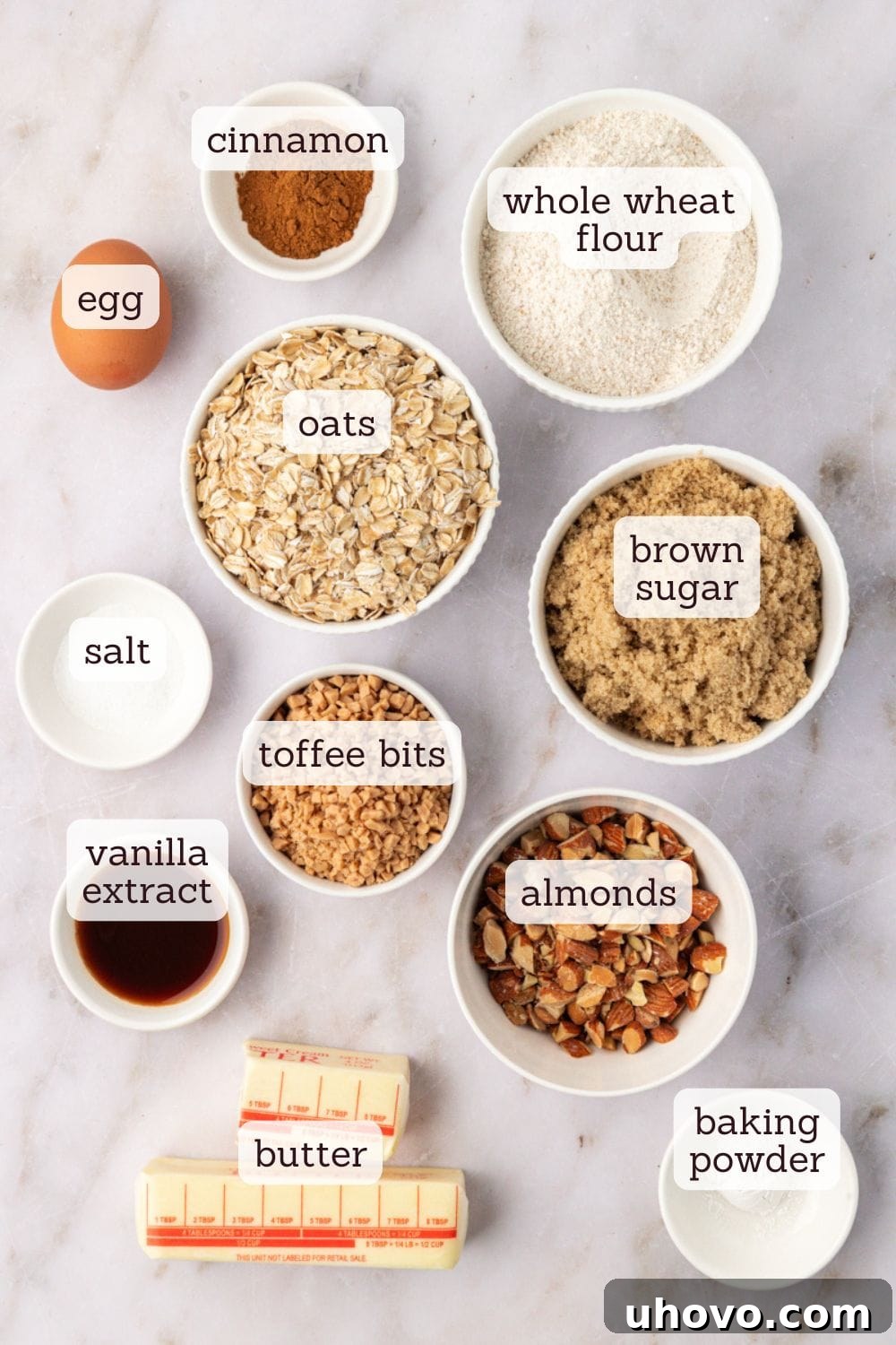 Overhead view of all ingredients for Toffee Almond Oatmeal Cookies laid out on a white surface, including flour, oats, butter, sugar, toffee bits, and almonds.