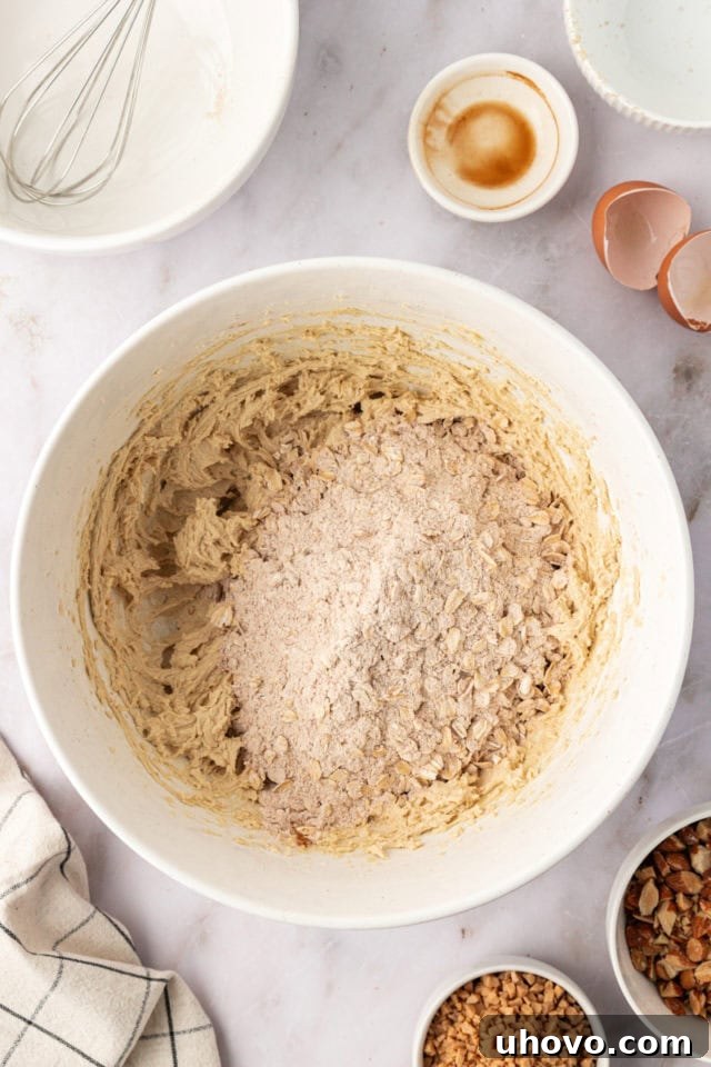 Overhead view of dry ingredients being gradually added to the wet ingredients in a mixing bowl for toffee almond oatmeal cookies.