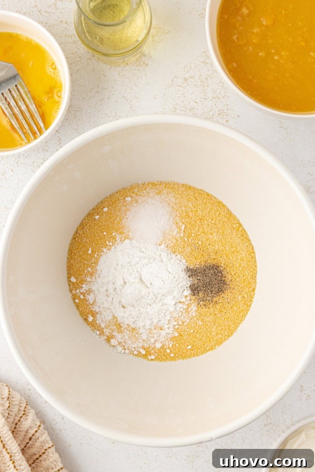 Overhead view of cornmeal, baking powder, salt, and pepper measured out in a white mixing bowl, ready to be combined.
