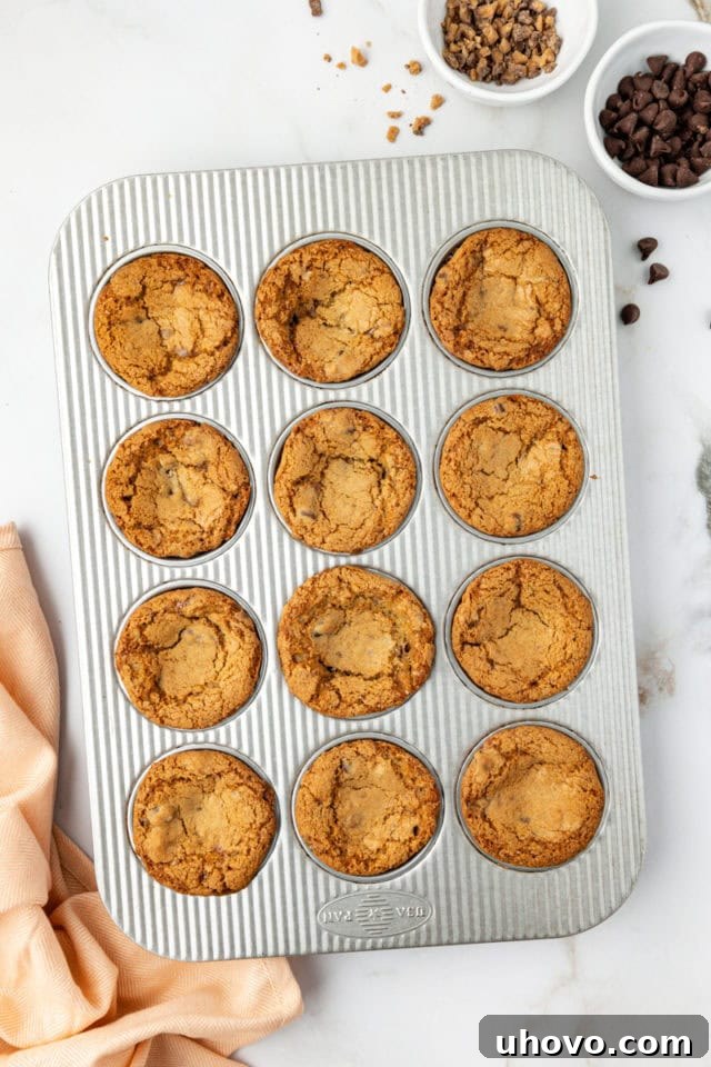 Overhead view of a muffin pan freshly pulled from the oven, containing 12 golden-brown blondie cupcakes with slightly sunken centers, indicating they are perfectly baked.