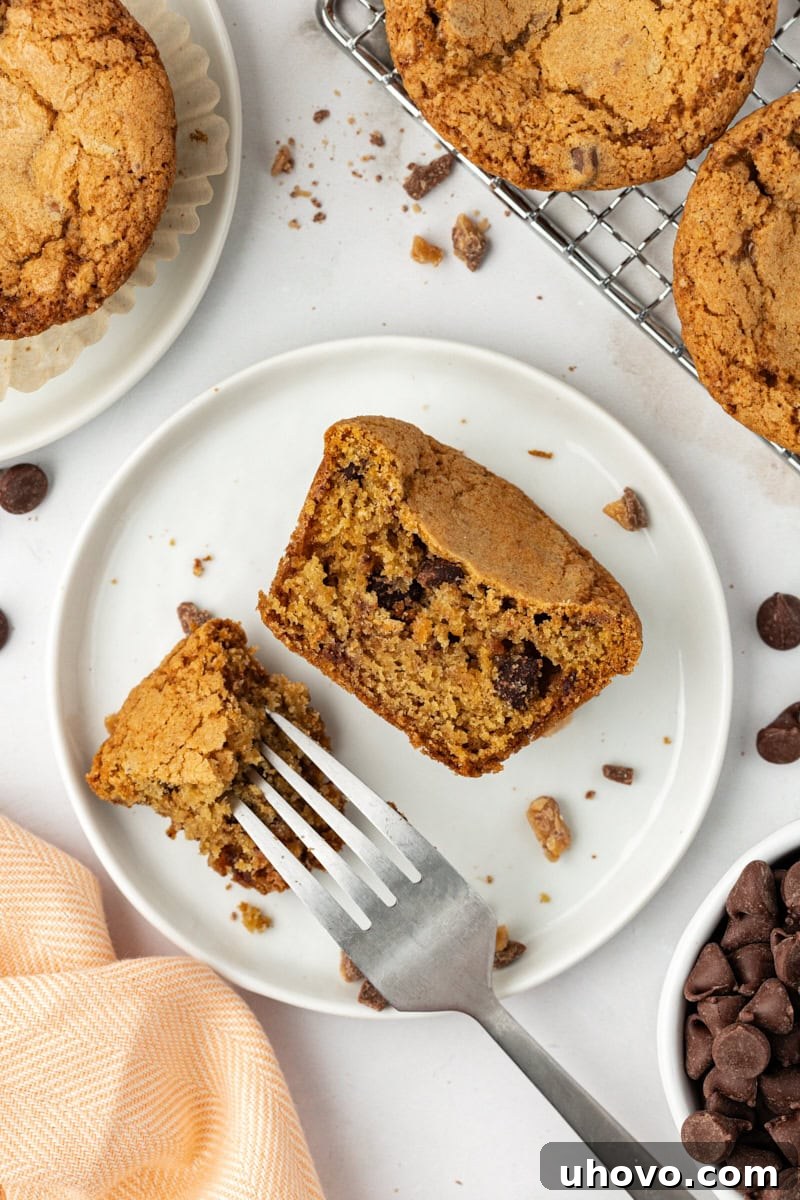A delectable close-up of half a blondie cupcake, showcasing its perfectly baked, chewy interior packed with chocolate chips and toffee bits. It rests on a white plate with a bite taken by a fork, surrounded by more whole blondie cupcakes, inviting a taste.
