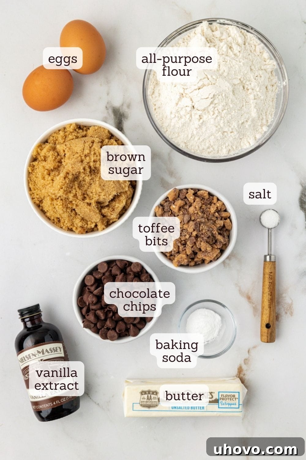 An overhead view showcasing the essential ingredients for blondie cupcakes neatly arranged on a countertop: flour, baking soda, salt, softened butter, brown sugar, eggs, vanilla extract, semisweet chocolate chips, and toffee bits.