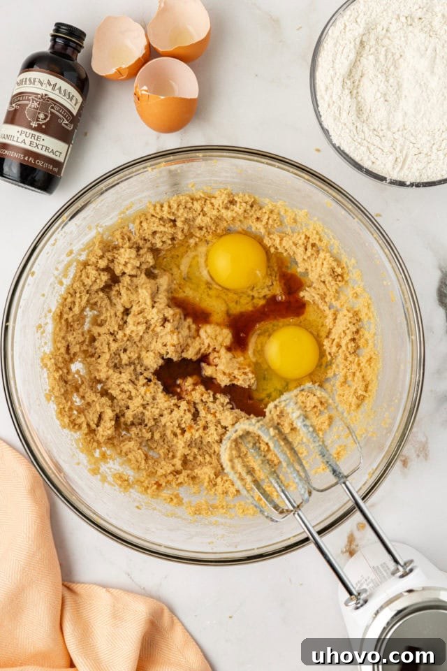 Overhead view of two large room-temperature eggs and a stream of vanilla extract being added to the creamed butter and brown sugar mixture in a glass mixing bowl, ready for incorporation.