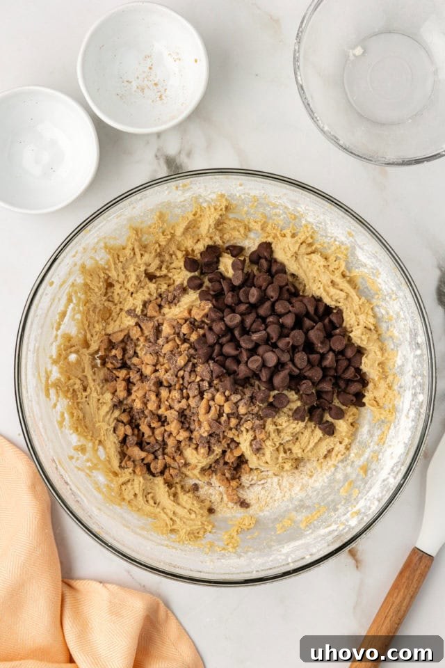 Overhead view of semisweet chocolate chips and buttery toffee bits being folded into the golden blondie batter in a glass mixing bowl, distributing the delicious add-ins.