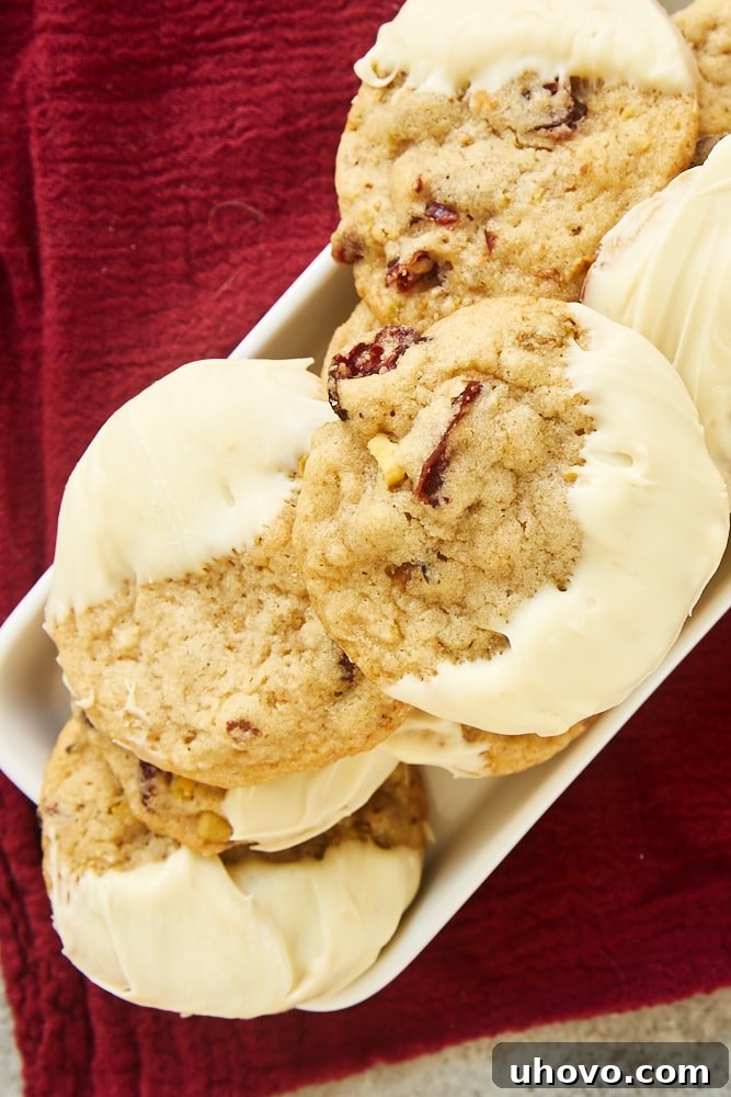 Top-down view of beautifully arranged Cranberry Pistachio Cookies, some dipped in white chocolate, presented on a small white tray against a rich dark red fabric.