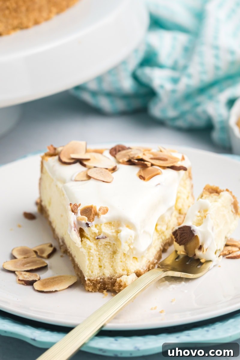 A perfectly portioned slice of creamy Amaretto Cheesecake, topped with a glossy white sour cream layer and golden toasted almond slivers. A fork has just taken a bite, revealing the smooth, rich interior and crumbly golden crust.