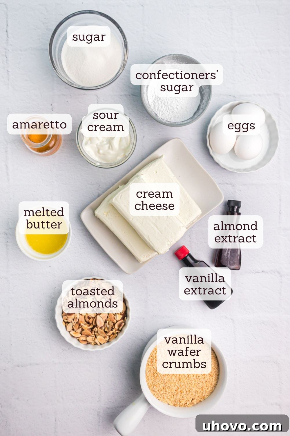An overhead shot of all the key ingredients neatly arranged on a white surface, including blocks of cream cheese, sugar, eggs, bottles of amaretto and extracts, sour cream, and vanilla wafers for the crust.