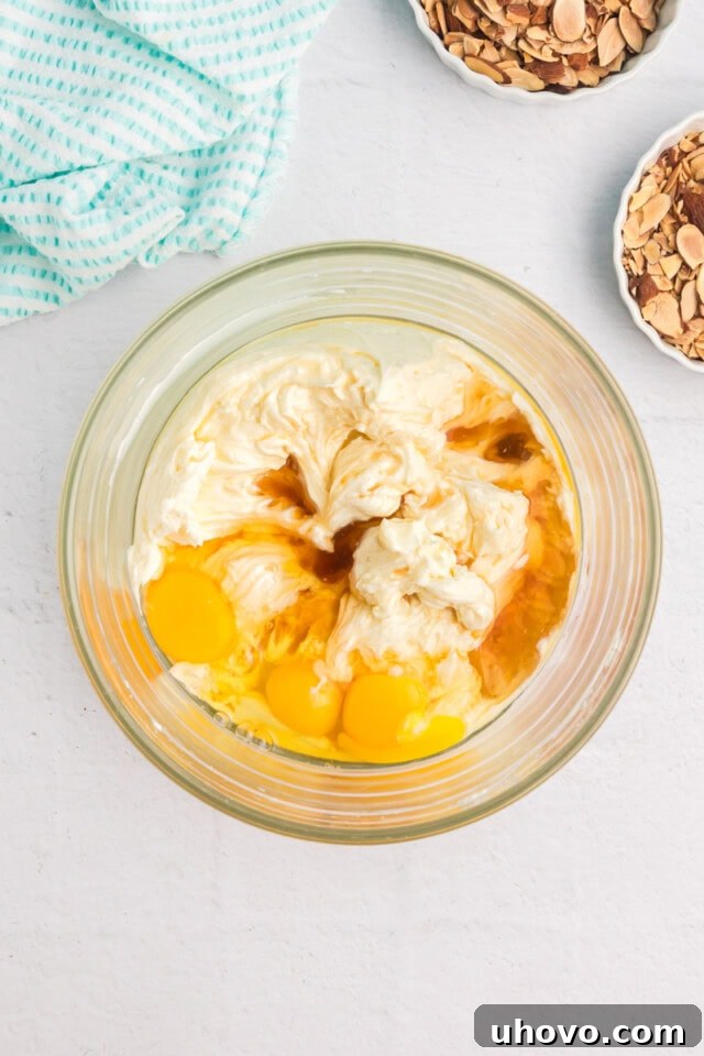 Overhead view of the creamy mixture of beaten cream cheese and sugar in a glass bowl, with eggs, amaretto liqueur, and vanilla and almond extracts carefully added, ready for gentle incorporation.