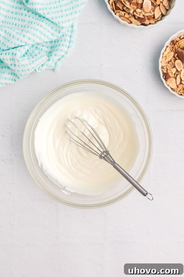 Overhead view of a small, clear glass bowl containing a smooth, creamy sour cream glaze, ready to be spread over the cooled cheesecake.