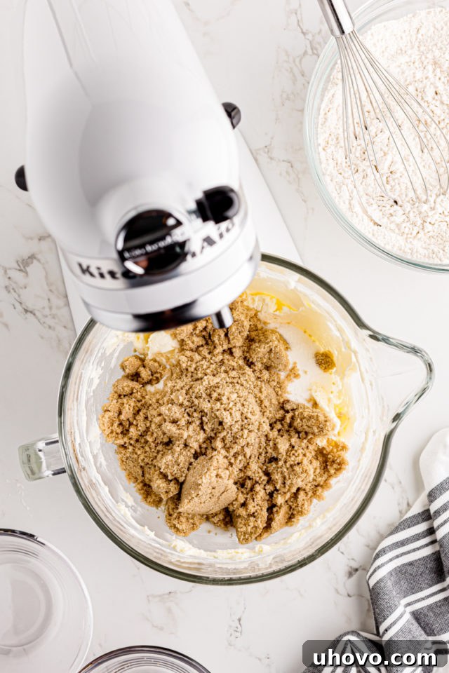 Granulated sugar and light brown sugar being added to the creamy mixture of butter and cream cheese in a stand mixer.
