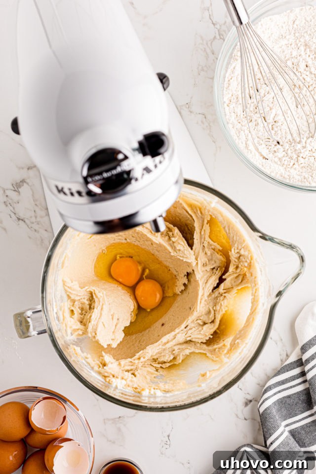 Room temperature eggs being carefully added one by one to the creamed butter, cream cheese, and sugar mixture, followed by vanilla extract.