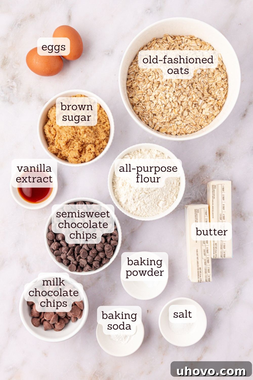 Overhead view of various key ingredients for brown sugar oatmeal chocolate chip cookies arranged on a white surface, including oats, flour, chocolate chips, and butter.