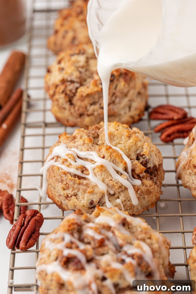 Close-up shot of a smooth, white glaze being drizzled over a warm cinnamon bun scone resting on a wire rack, with other scones and pecan halves in the background.