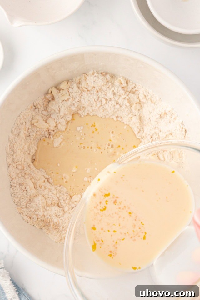 Overhead view of a liquid mixture (milk, egg, vanilla) being poured into the dry ingredients and butter mixture in a white mixing bowl.
